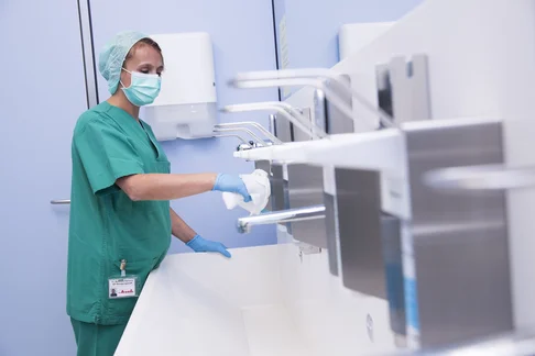 Eine Bild von einer Frau im grünen OP Kleidung mit Mundschutz die Seifen- und Desinfektionsspender wischt