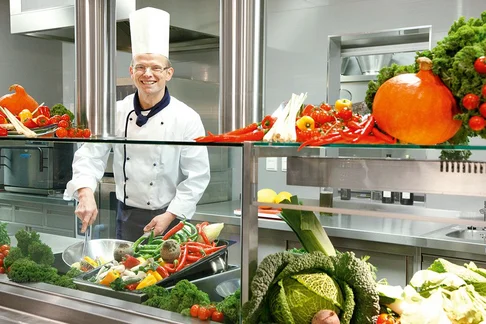 Ein Bild von einem Koch der lächelnd in der Küche steht und ein Gericht im Wok kocht, in der Küche steht überall frisches und buntes Gemüse