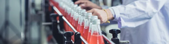 Ein Bild von Flaschen mit rotem inhalten auf dem Band einer Maschinellen Anlage, eine Person mit weißem Kittel steht neben dran